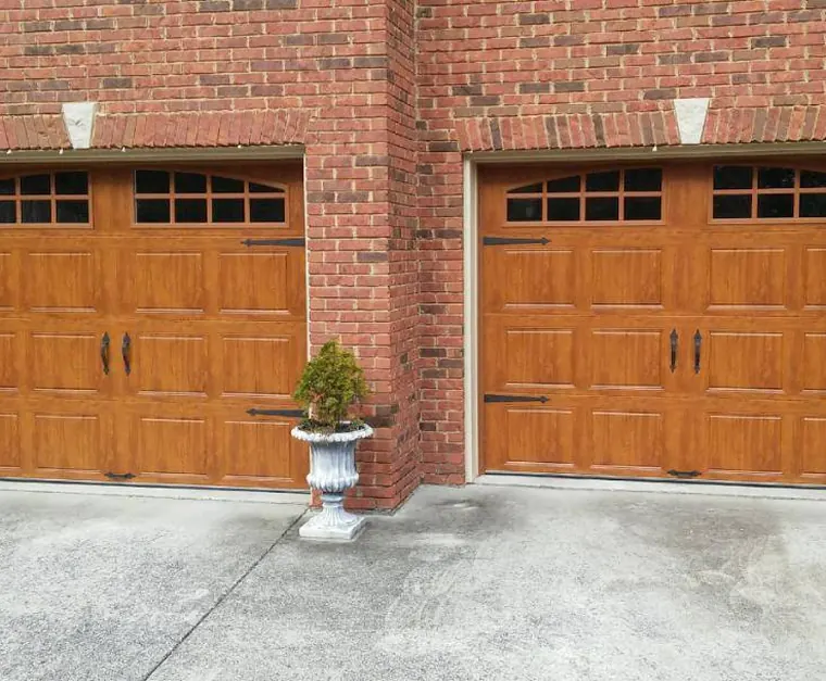 Two wood-grain carriage garage doors with arched windows and black hardware on a red brick house
