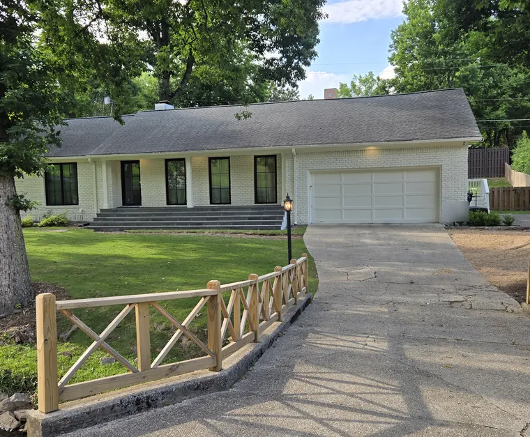 A white-painted brick ranch house with a white two-car garage door, a long driveway and a wooden X-rail fence