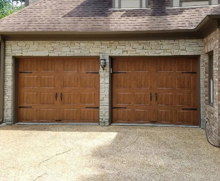 Two wood-tone carriage garage doors with black hardware on a stone and brick house