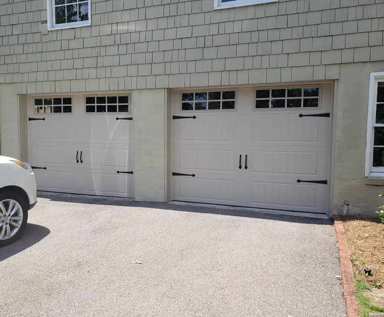 Two tan carriage-style garage doors with window grids and black hinges on a shingle-sided house