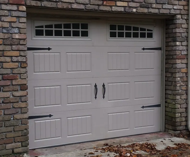 A tan carriage-style garage door with arched windows and black hardware set in old brick, a wooden gate beside it