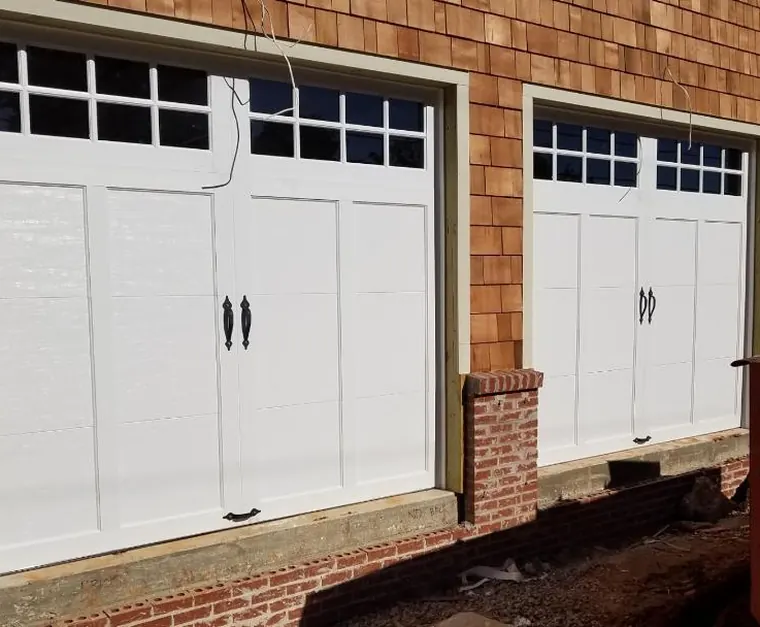 White carriage-style garage doors with window grids and black handles on a cedar-shingle house