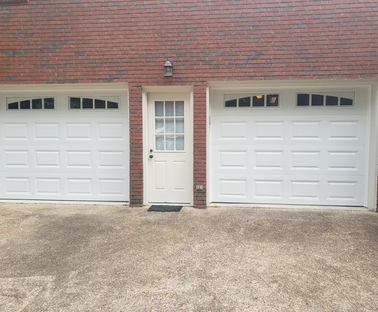 Two white raised-panel garage doors with arched window rows on a red brick house, a side door between them