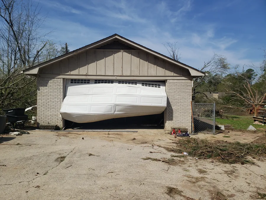 A white garage door buckled and bowed out of its opening, branches and debris on the driveway