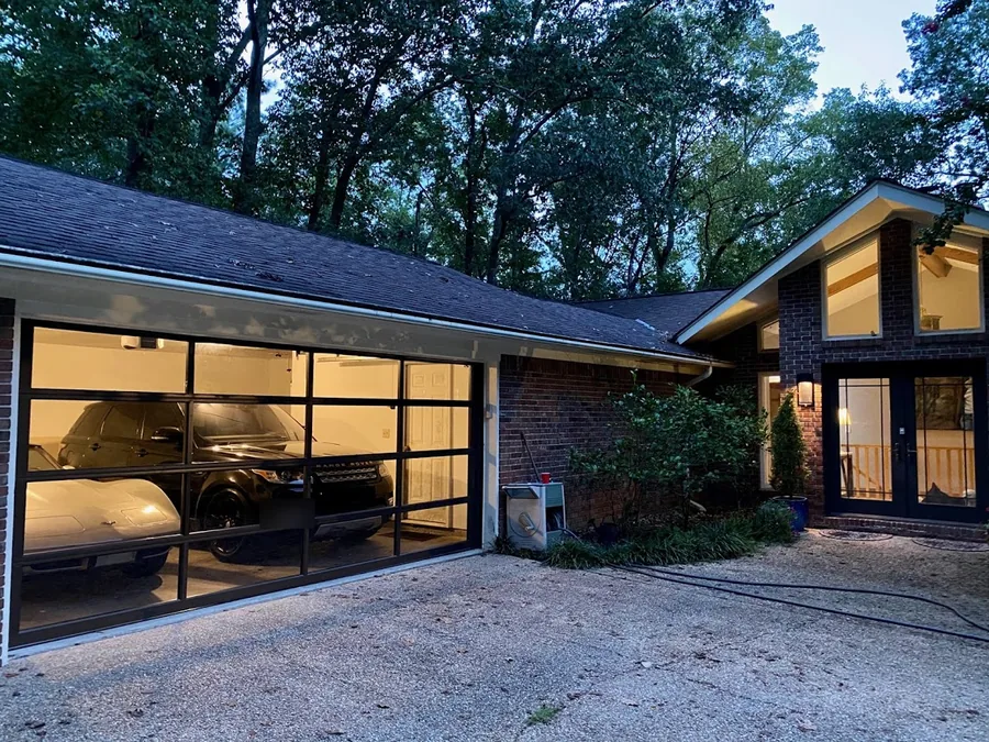 Full-view glass and black aluminum garage doors lit from inside at dusk