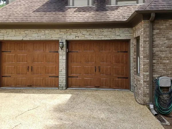 Two wood-tone carriage doors with black hardware on a stone and brick house