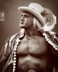 Sepia portrait of a man in a white feathered cowboy hat and a bead necklace, wearing a championship belt