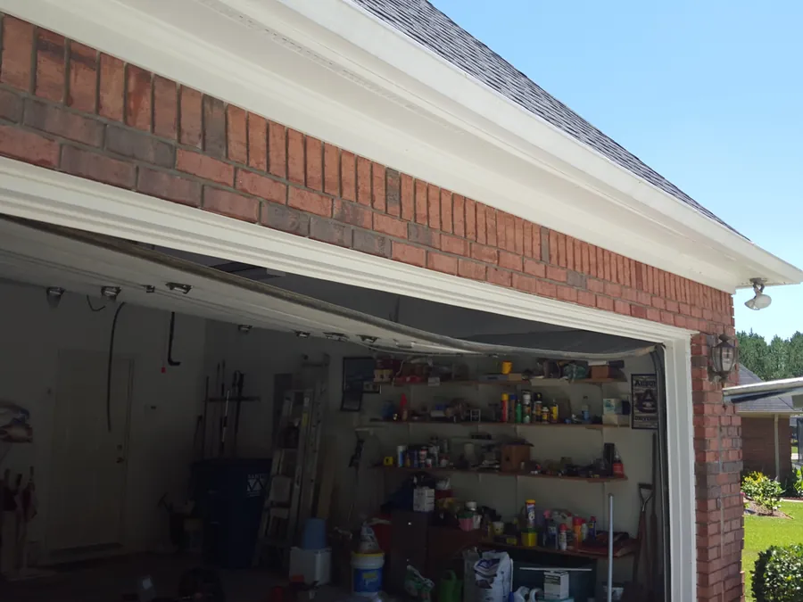 A garage door sagging out of shape across its opening on a brick house, the top section hanging loose