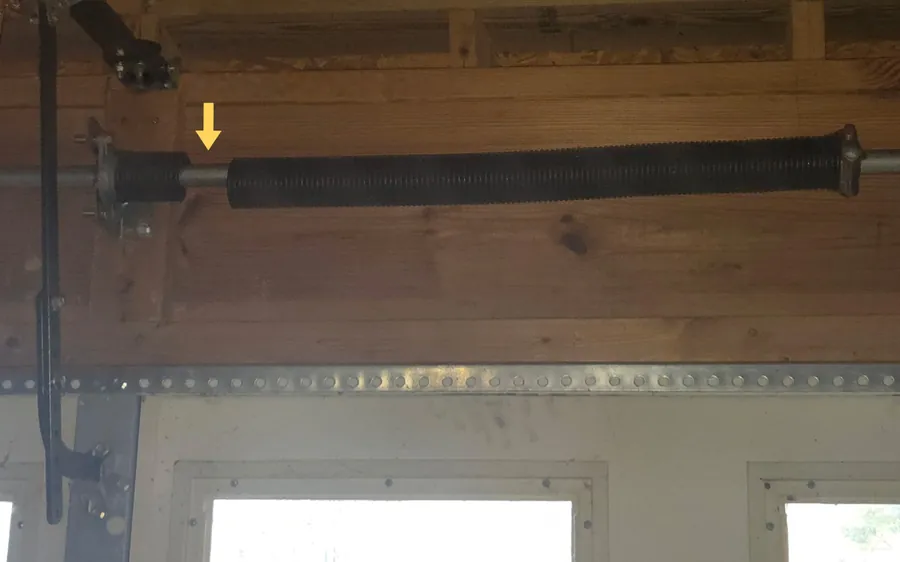 A torsion spring above a garage door, snapped near its left end, a yellow arrow pointing at the gap