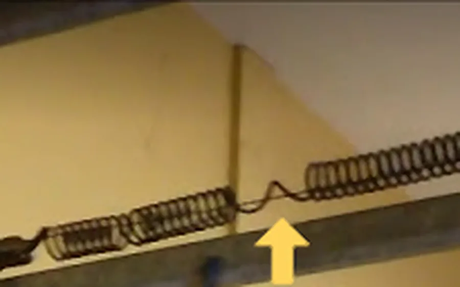 An extension spring along a garage door track, stretched open in the middle, a yellow arrow pointing at the gap